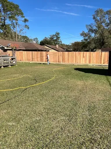 Lawn with new wooden fence, person standing in yard, trees, clear blue sky.