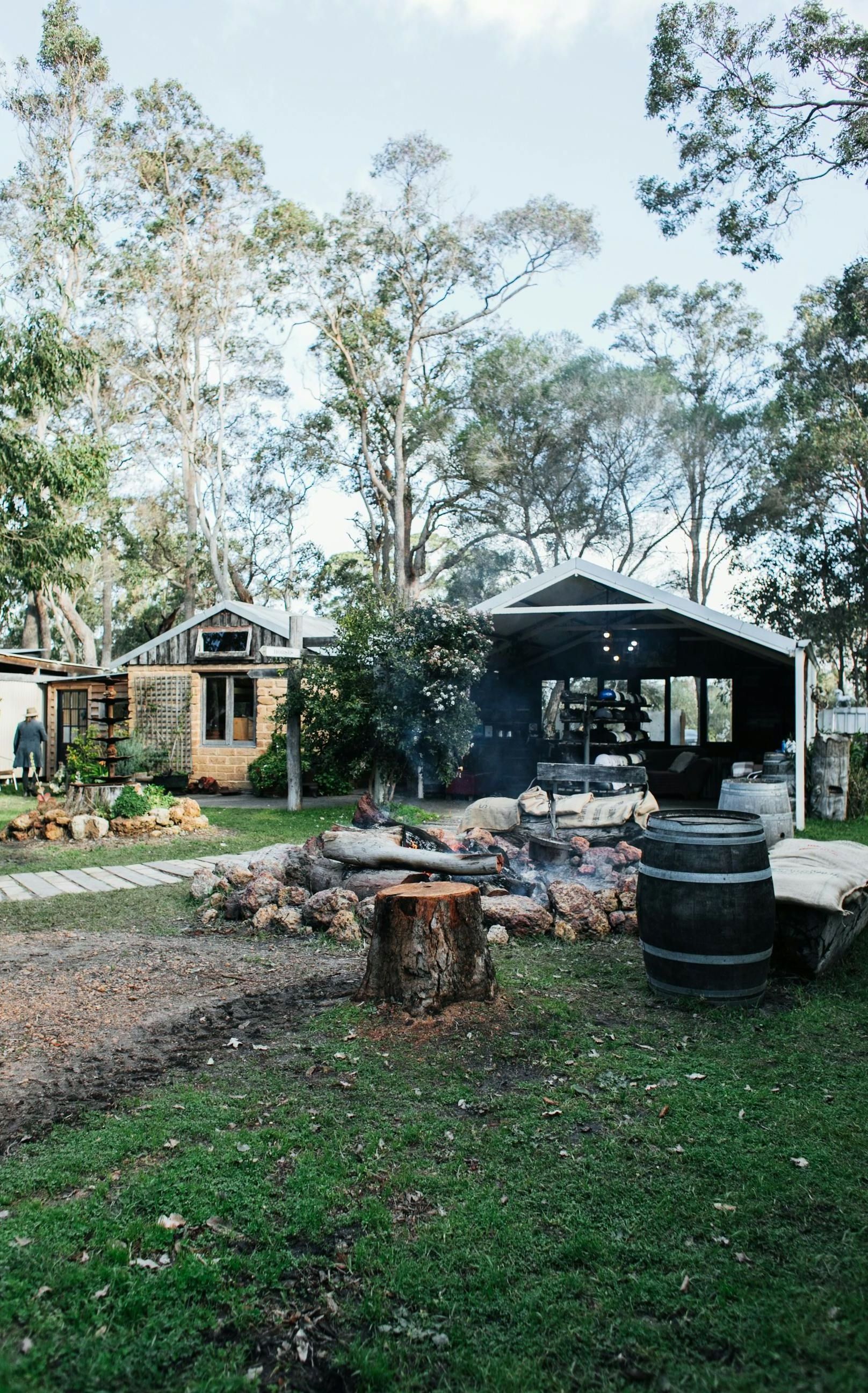 A rustic outdoor scene with a small cabin, an outdoor covered area, and a fire pit with a tree stump in front.