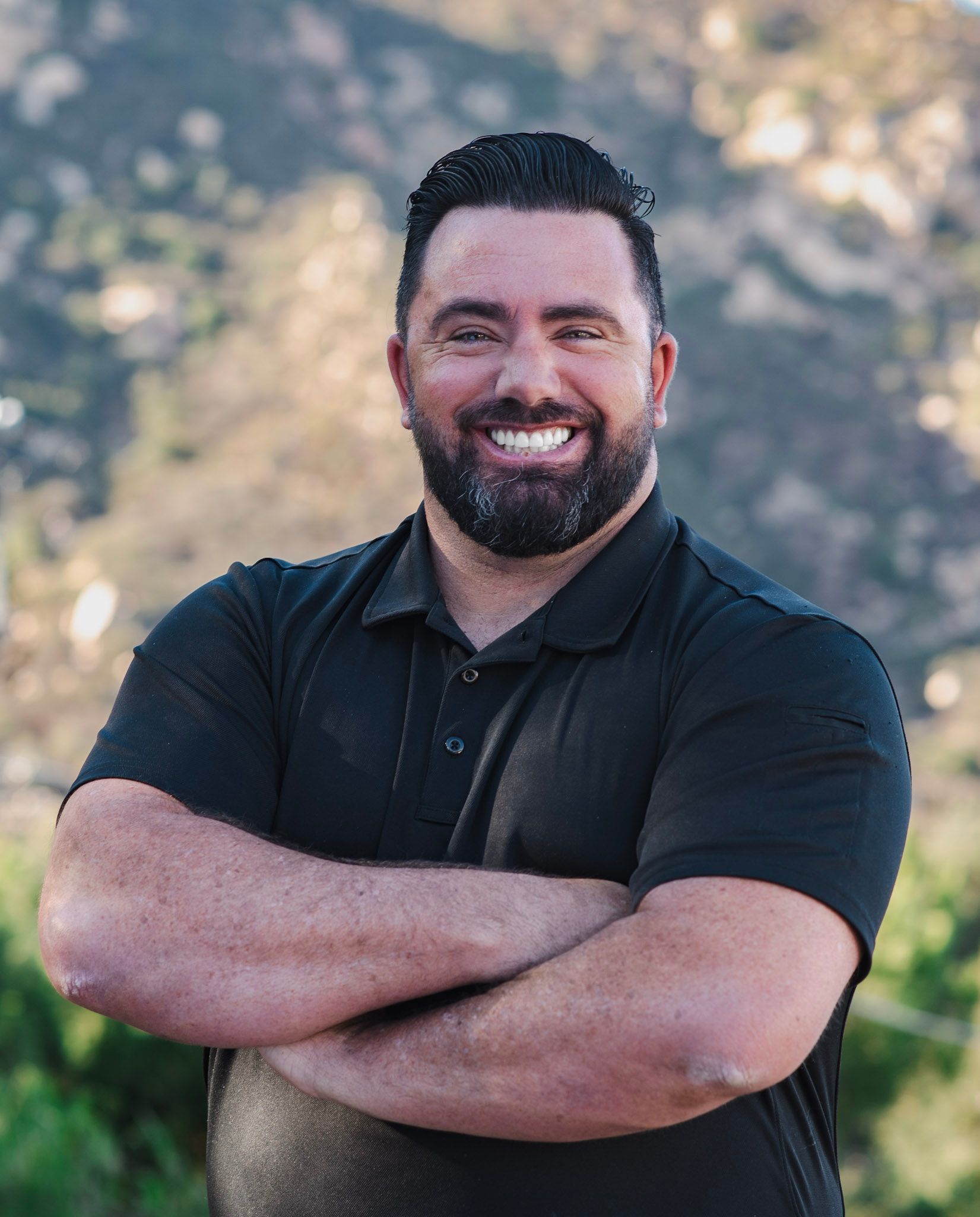 A man with a beard is smiling with his arms crossed in front of a mountain.