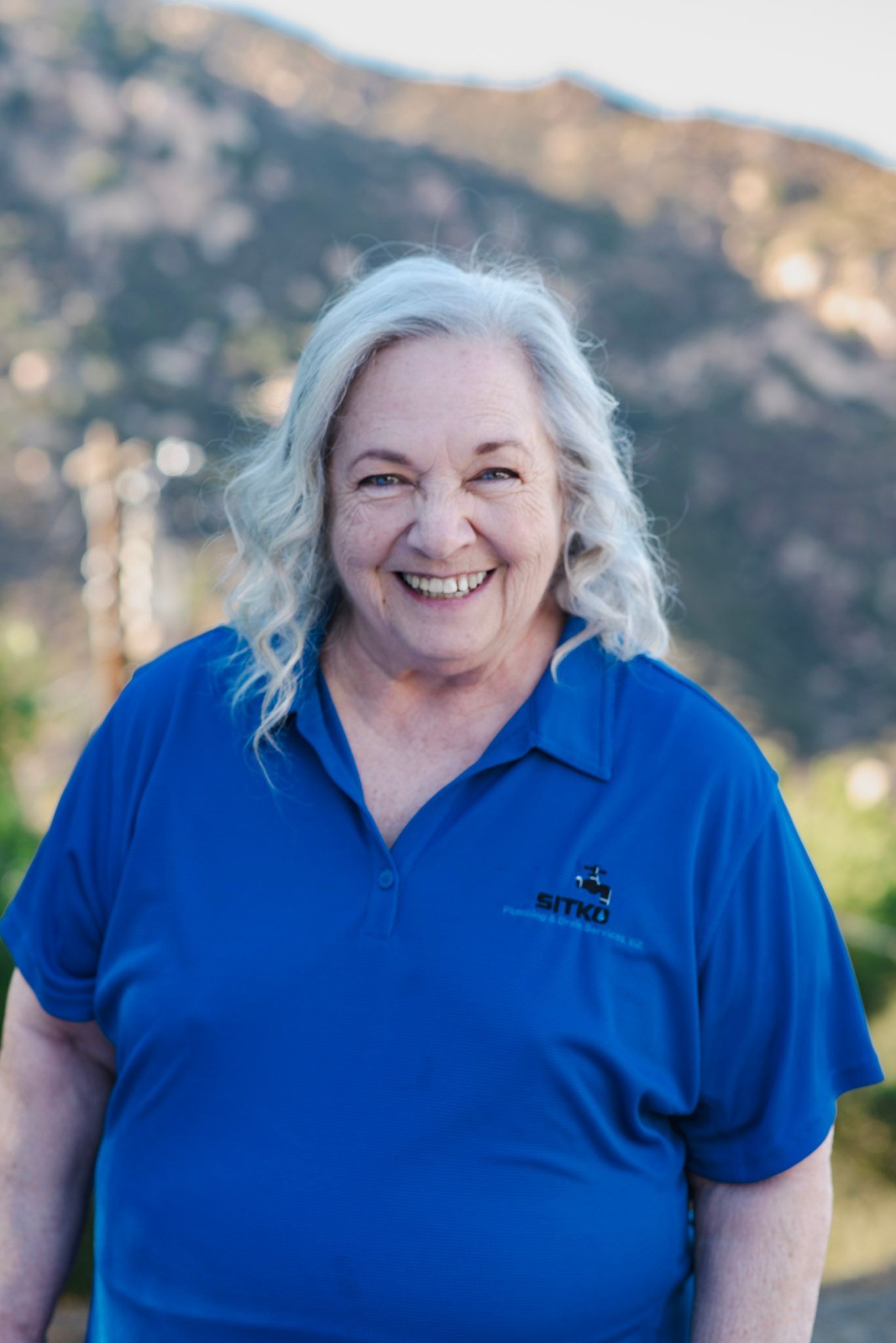 A woman in a blue shirt is smiling in front of a mountain.