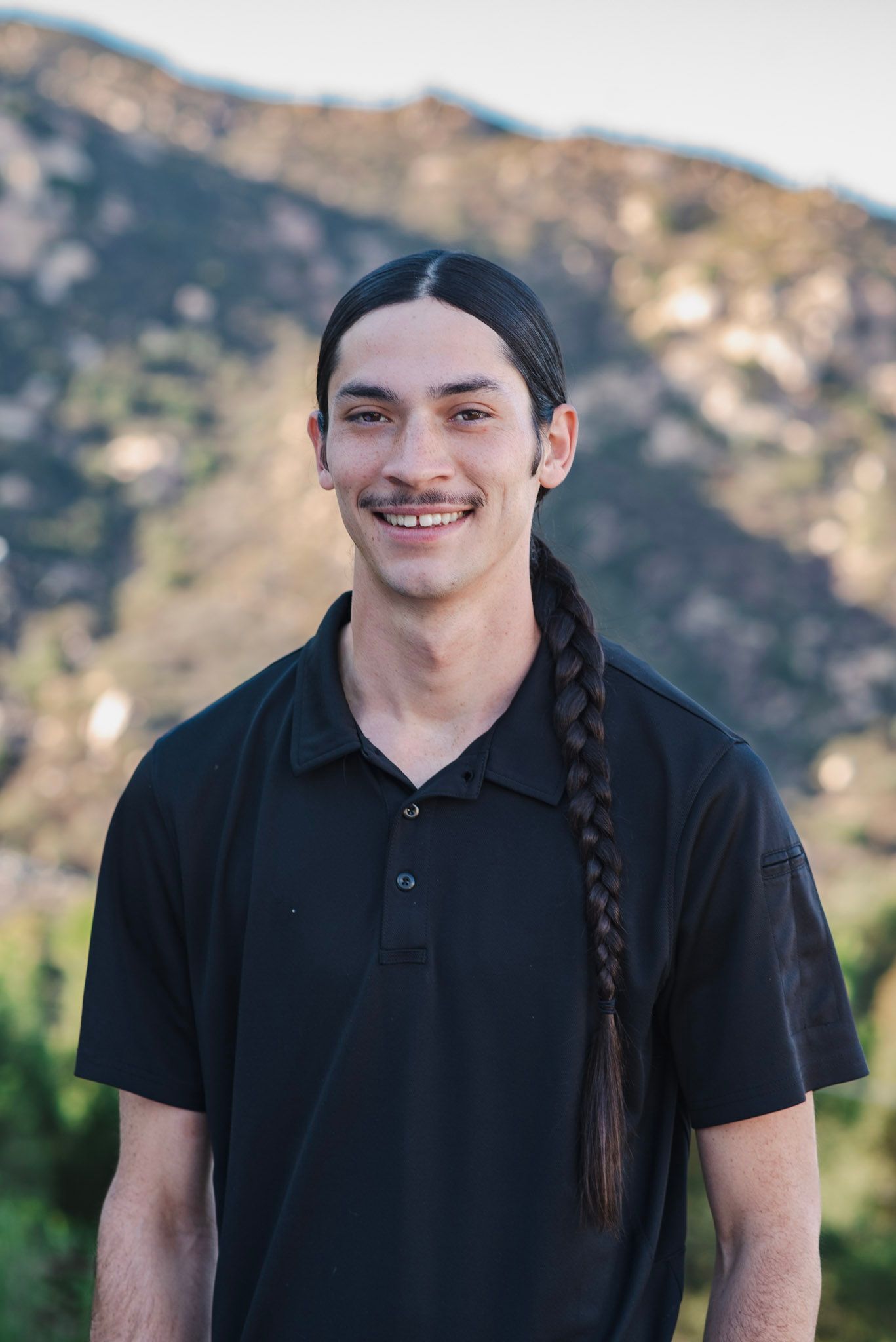 A young man with long hair is wearing a black shirt and smiling in front of a mountain.