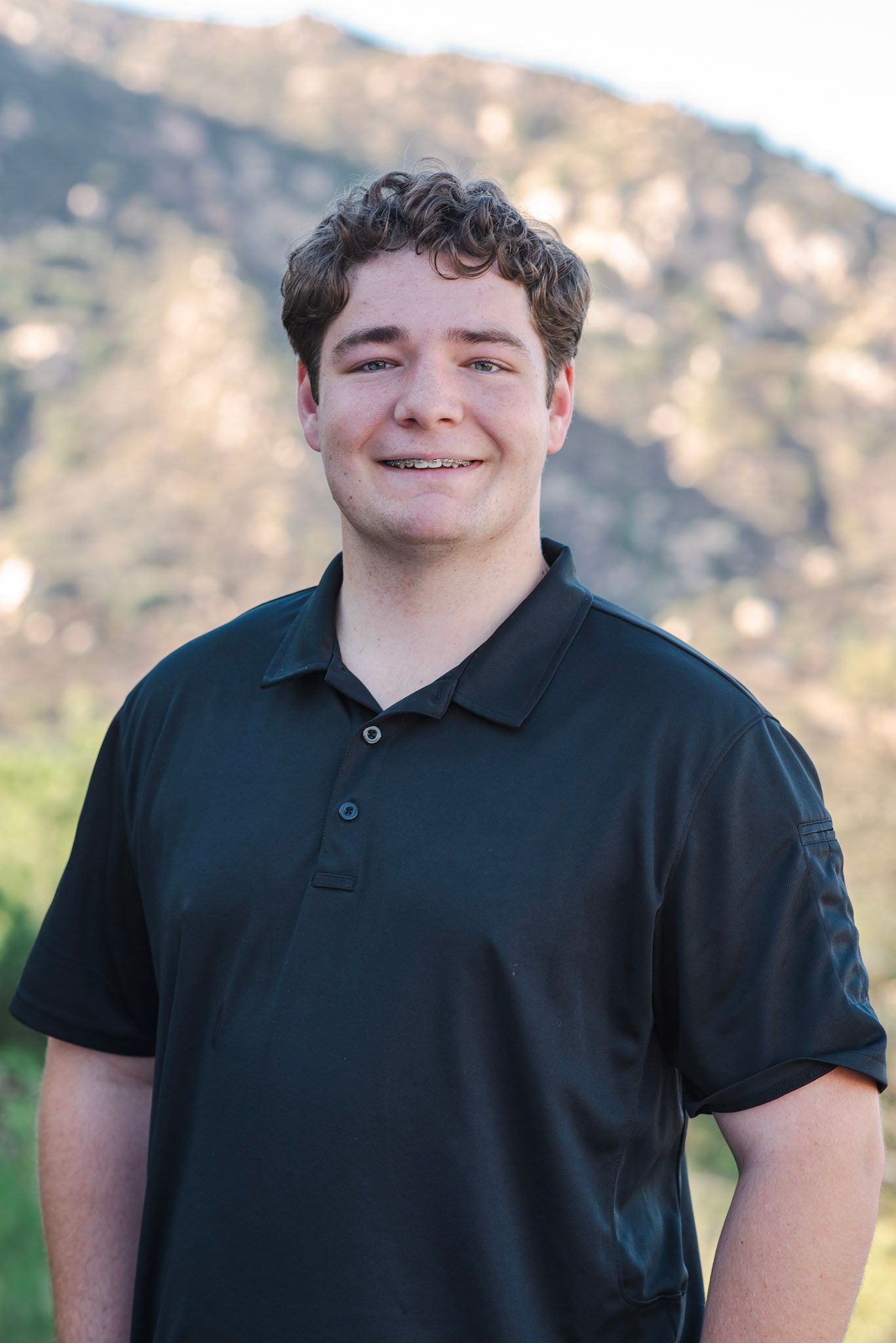 A young man in a black polo shirt is standing in front of a mountain.