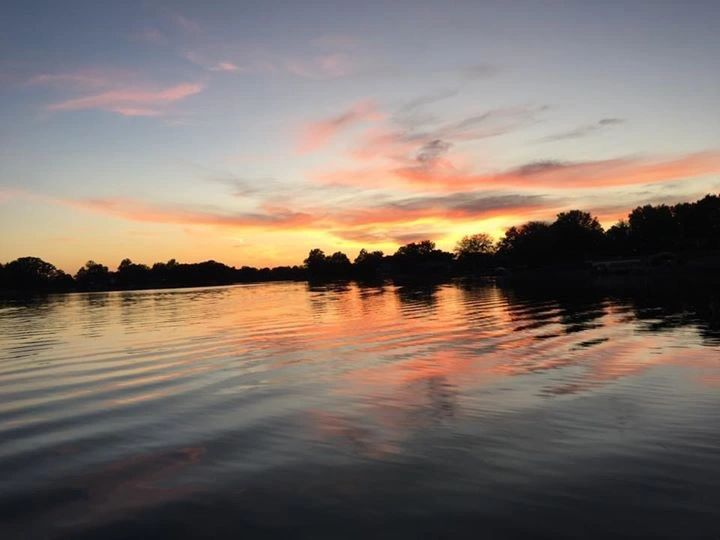 Sunset over calm lake, reflecting orange and pink clouds. Silhouetted trees on the horizon.