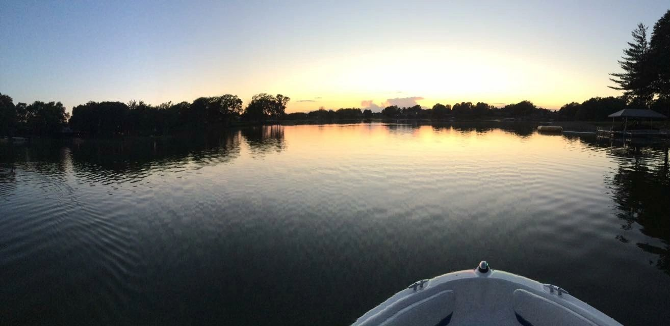 Boat on a calm lake reflecting a colorful sunset. Trees line the far shore.