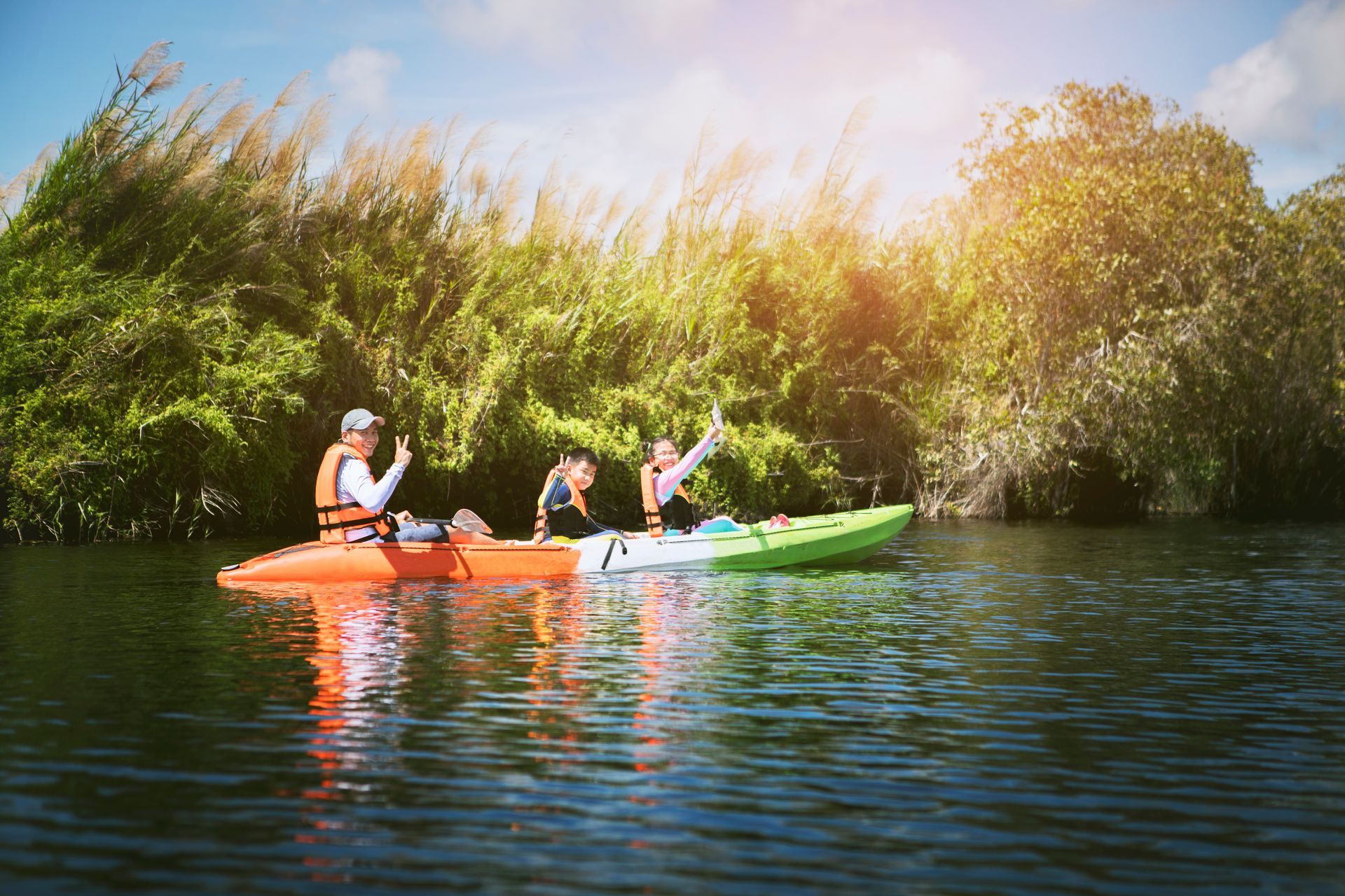 People kayaking on a calm river; lush green plants line the banks, bright sunlight.