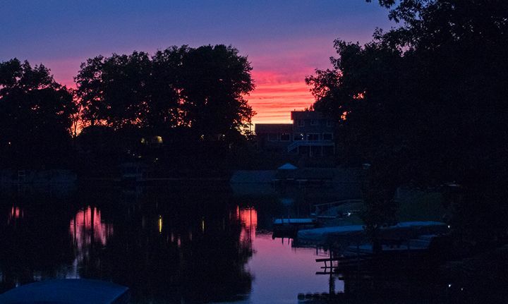 Sunset over a calm lake, reflecting vibrant pink and orange hues. Silhouetted trees and docks frame the water.