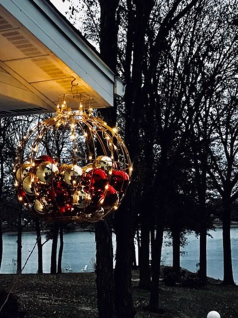 Ornate hanging Christmas ornament decoration with gold and red balls, lit with fairy lights, by a lake.