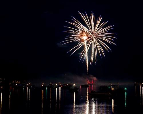 Fireworks burst over water at night; lights reflect.