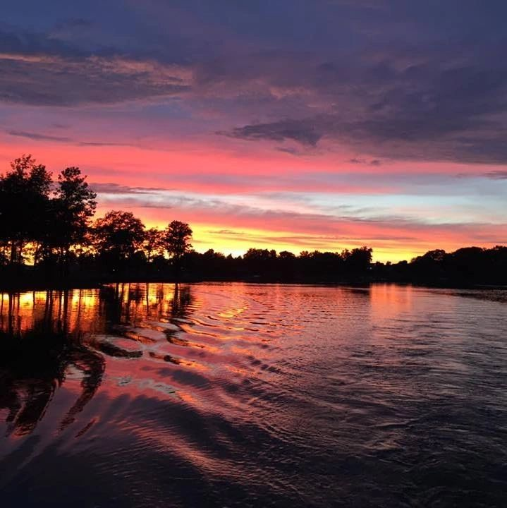 Sunset over a lake; orange, pink, and purple sky reflects in the water, silhouettes of trees on the shore.