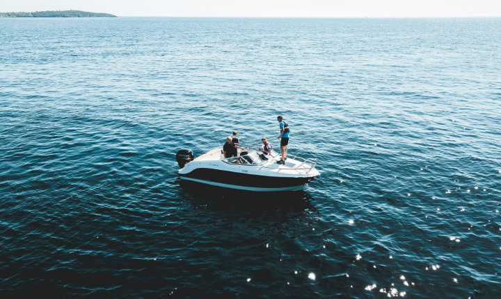 Boat with people on the water; a person stands at the back, others sit on deck. Blue water, sunny day.
