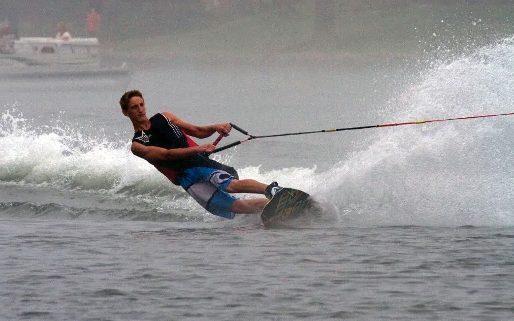 Man waterskiing, leaning sideways, with water spraying. Lake setting, overcast.