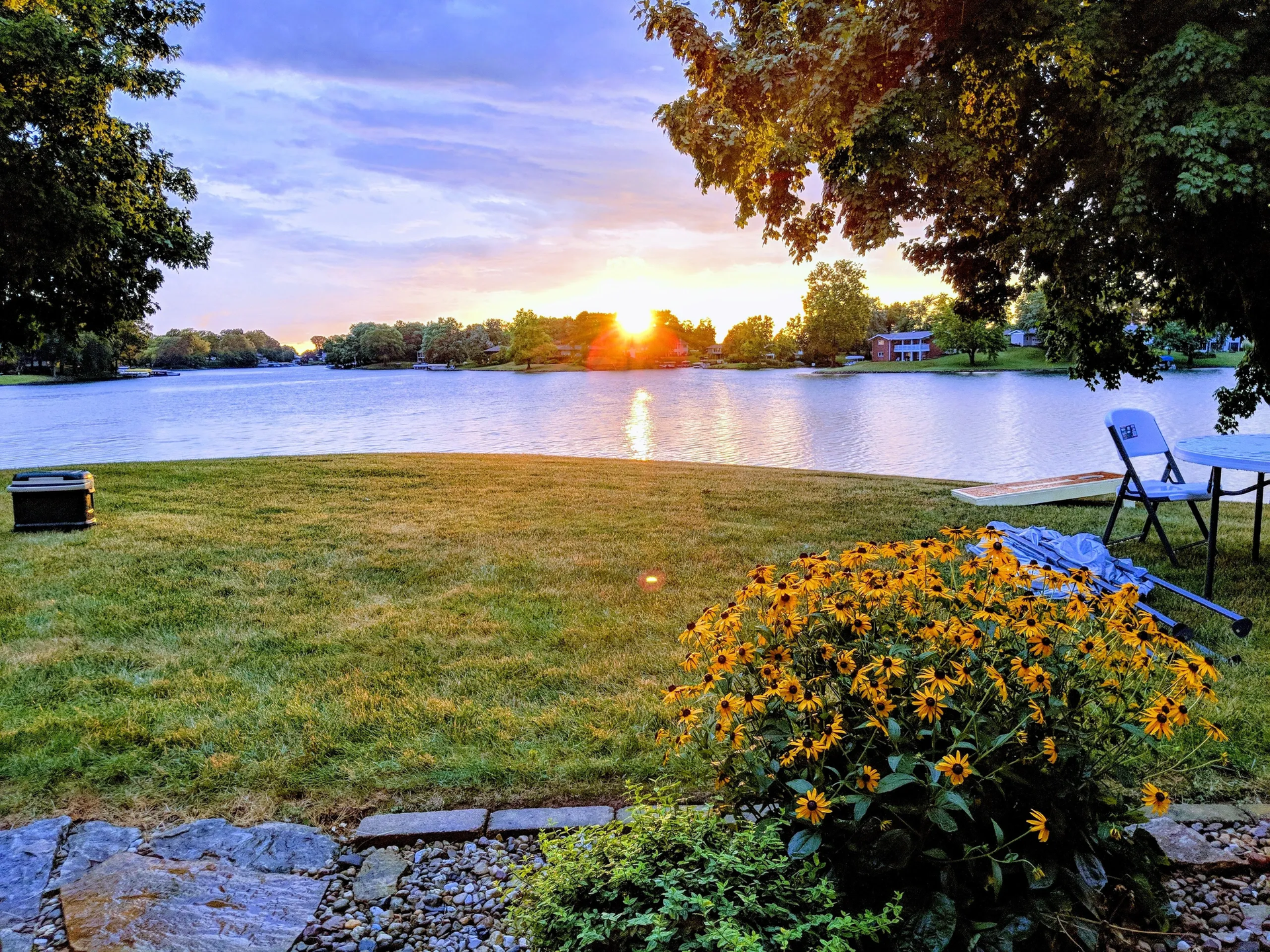 Sunset over a lake; golden light, green grass, yellow flowers, trees frame the view.