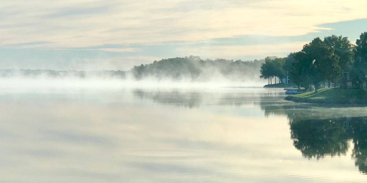 Mist hangs over a calm lake, reflecting the hazy sky and a treed shoreline.