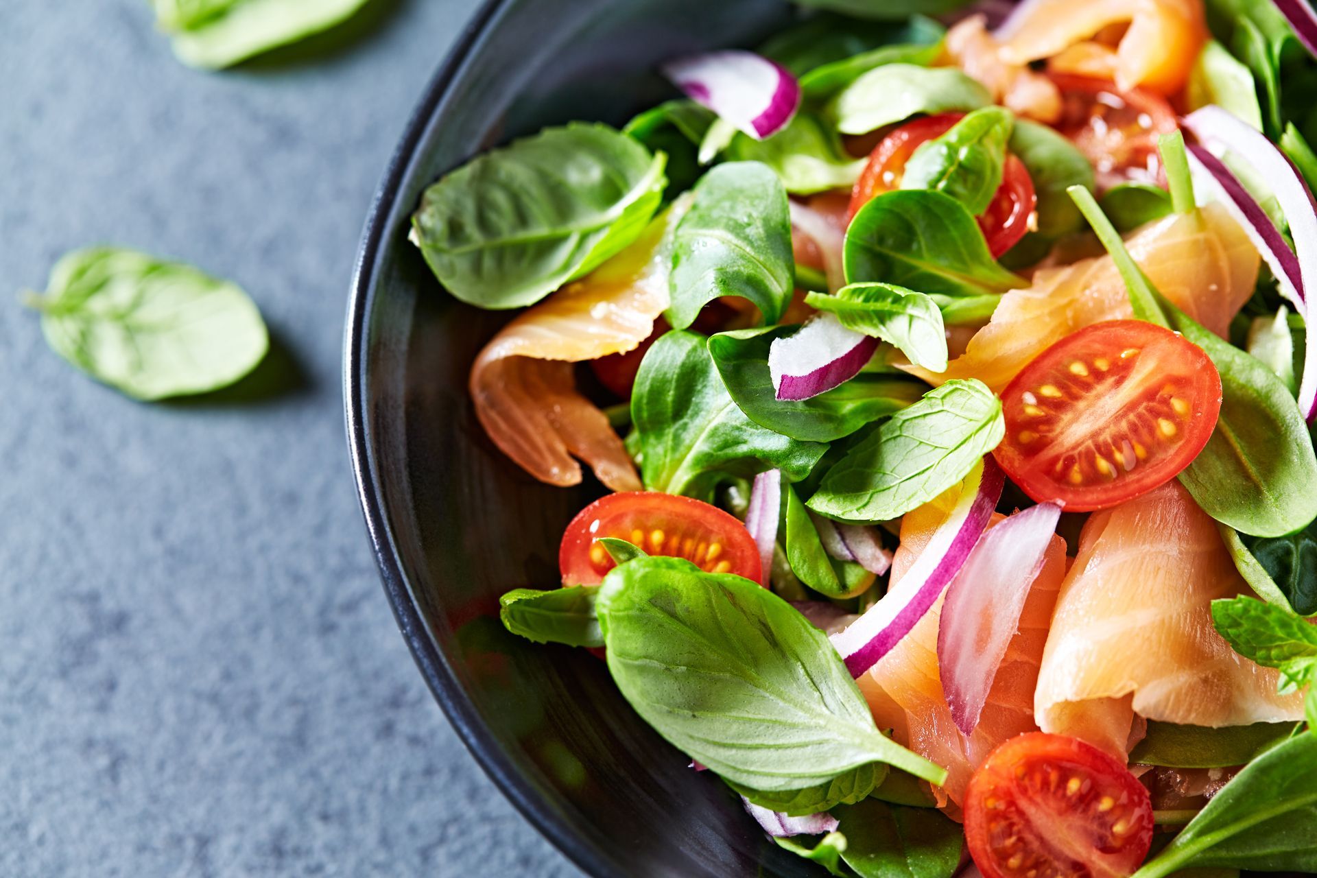 A salad with tomatoes , spinach , onions and salmon in a black bowl.