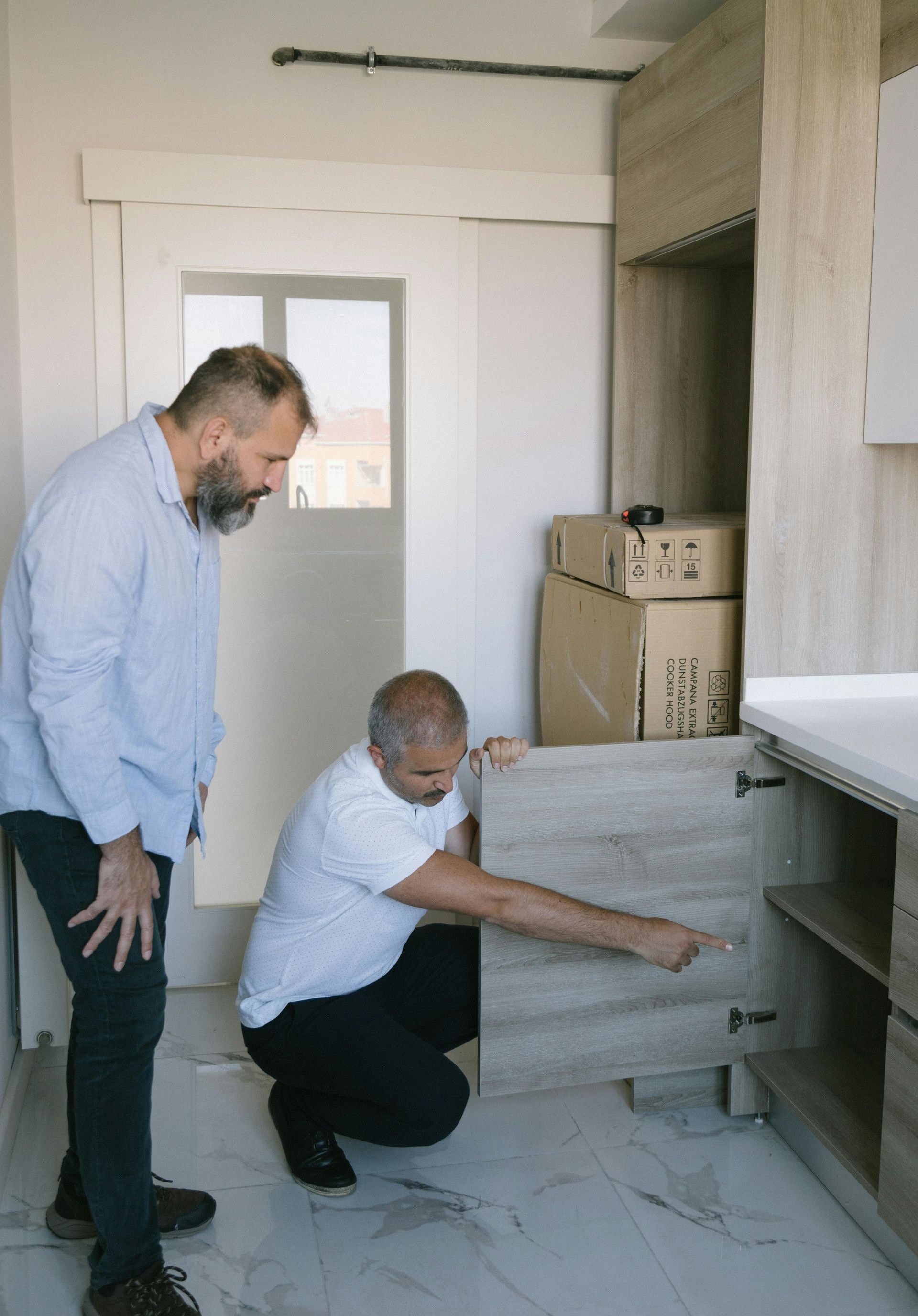 Two men are working on a cabinet in a kitchen.