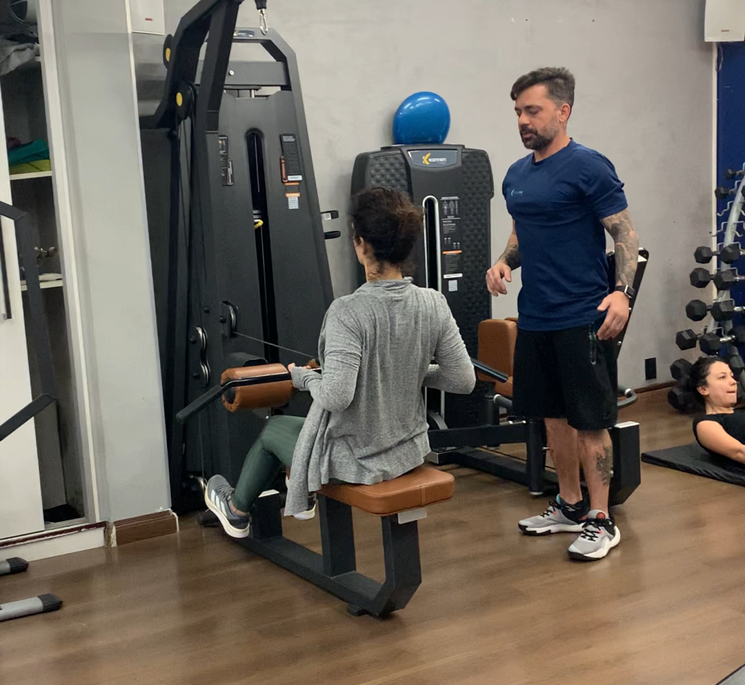 Um homem está de pé ao lado de uma mulher sentada em uma máquina em uma academia.