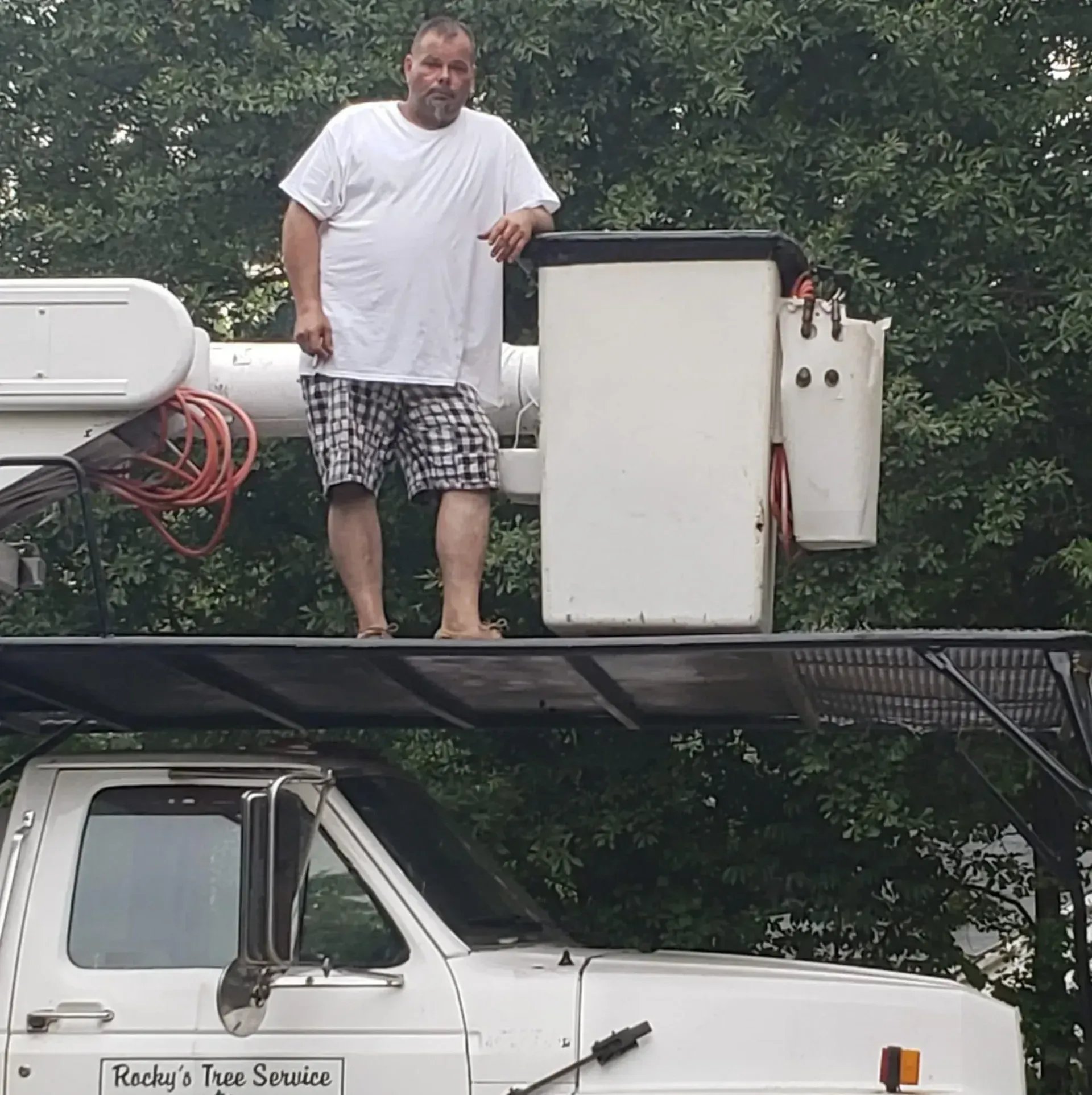 Man standing in the bucket of a white service truck, wearing a white t-shirt and plaid shorts. Trees in background.