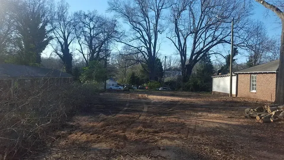 Cleared lot with brown earth, bordered by trees and houses under a blue sky.