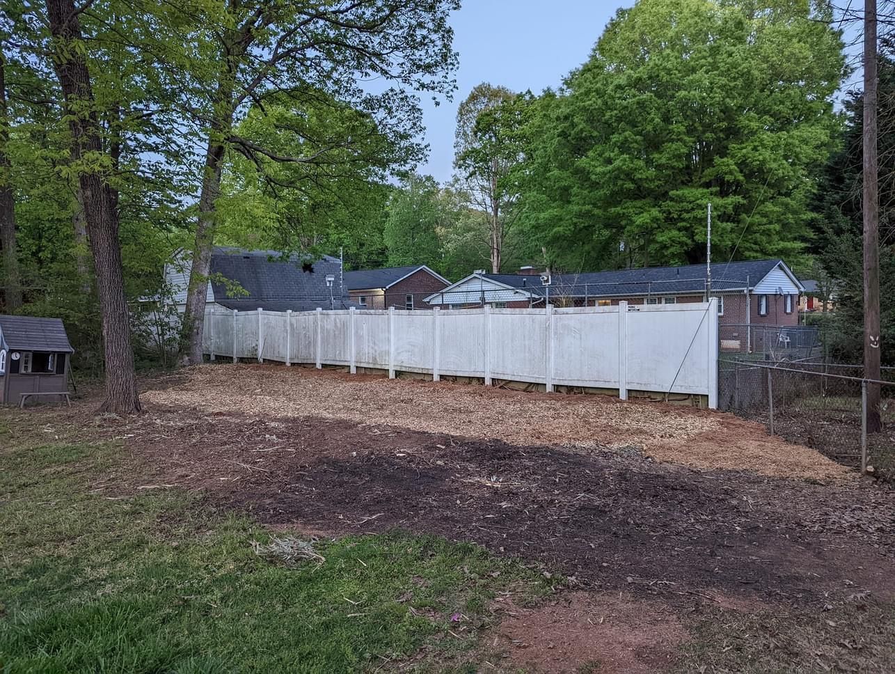 White fence bordering a yard with woodchips and trees. Buildings visible in the background.