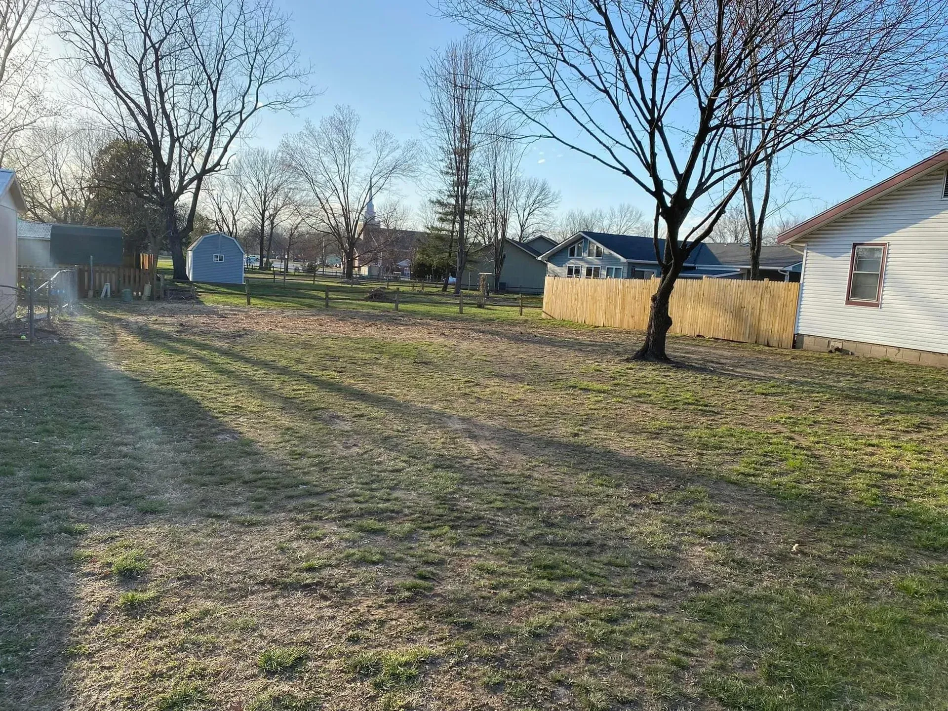 Grassy backyard with sparse trees, a fence, and houses in the distance on a sunny day.