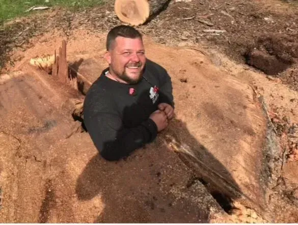 Man smiling, emerging from a hollowed-out tree trunk in a wooded setting.