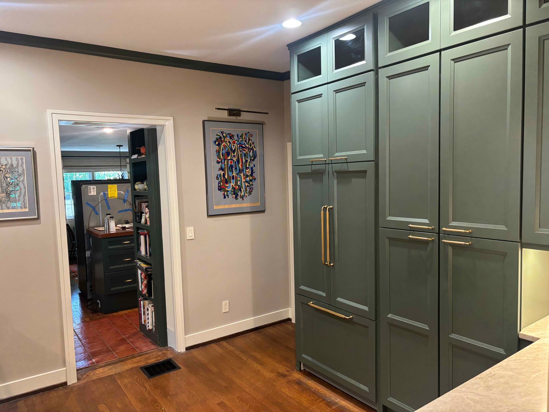 A kitchen features tall, olive-green cabinetry with gold hardware next to a doorway looking into an adjacent room.