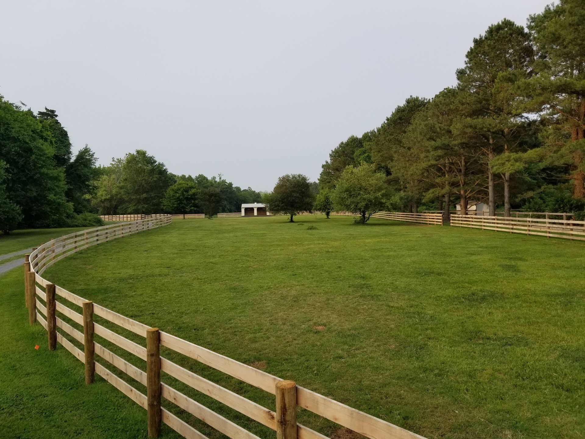 A split-rail fence curves around a green, grassy pasture leading toward trees and a small structure in the distance.