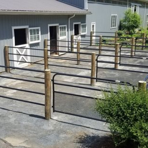 A series of horse stalls with black metal gates and wooden posts attached to a grey barn exterior.