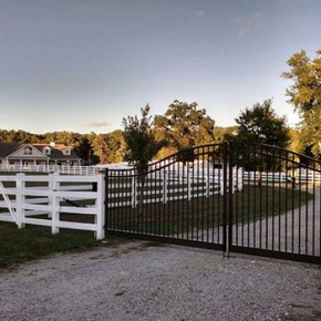 A black arched metal gate stands open on a gravel driveway, leading to a white-fenced rural property with a large house.