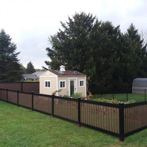 A small, tan shed with a roof cupola sits inside a black metal and wood-slatted fenced garden on a green lawn.
