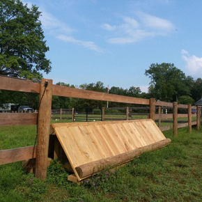 A wooden equestrian cross-country jump sits in a grassy field in front of a wooden split-rail fence under a blue sky.