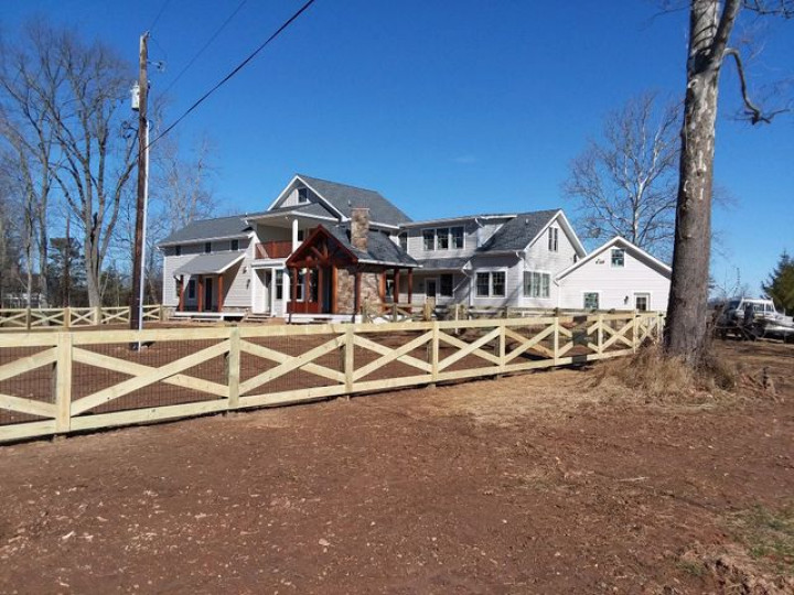 A large white farmhouse with gray roofing and a rustic wood porch sits behind a long, wooden X-pattern fence.