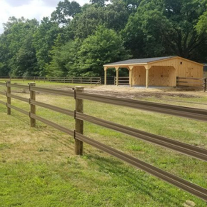A wooden three-rail fence runs through a grassy field toward a wooden run-in shed near a tree line.