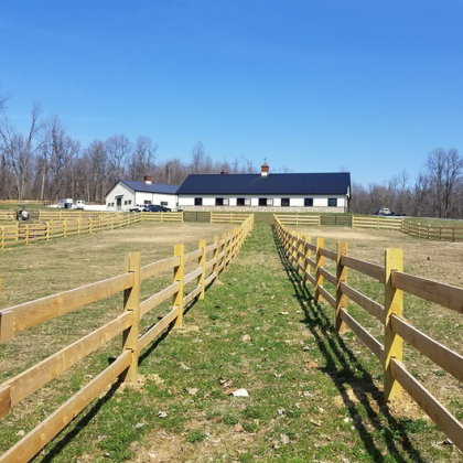 A path leads between two wooden fences toward a long, white barn with a dark roof under a clear blue sky.