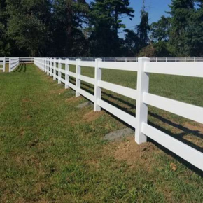 A white three-rail vinyl fence stretches across a green grassy field under a clear blue sky.