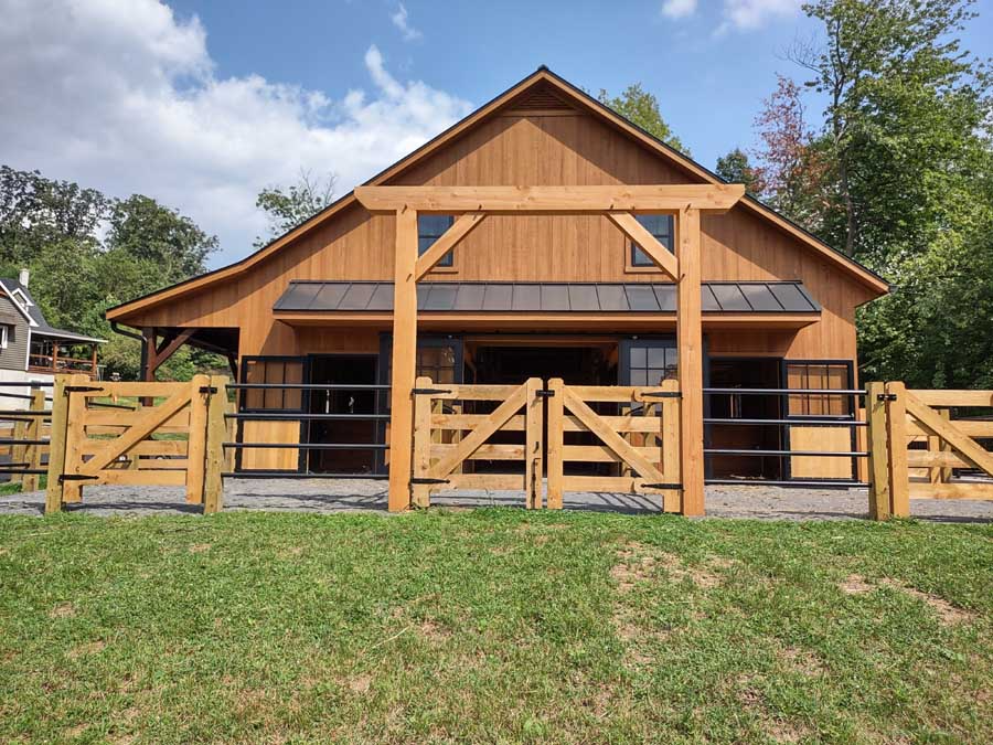 A rustic wooden horse barn featuring a gated entrance, metal roof, and surrounding fenced paddock under a blue sky.
