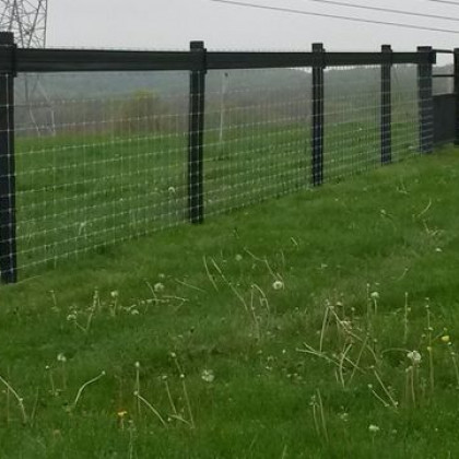 A black wooden post-and-wire farm fence stretches across a green grassy field on an overcast day.