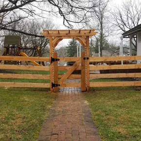 A wooden gate with an arbor topper sits centered in a split-rail fence, positioned over a brick path in a grassy yard.