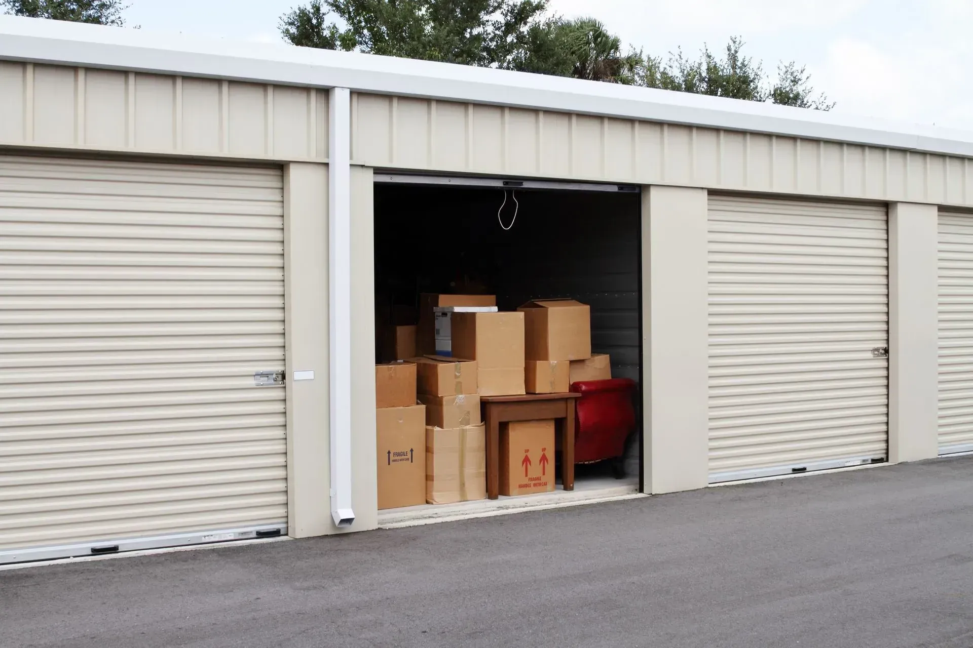 Storage unit with open door, filled with cardboard boxes and furniture. Building is beige.