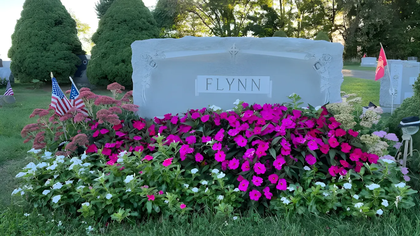 Stone memorial surrounded by bright pink and white flowers with small flags in a cemetery