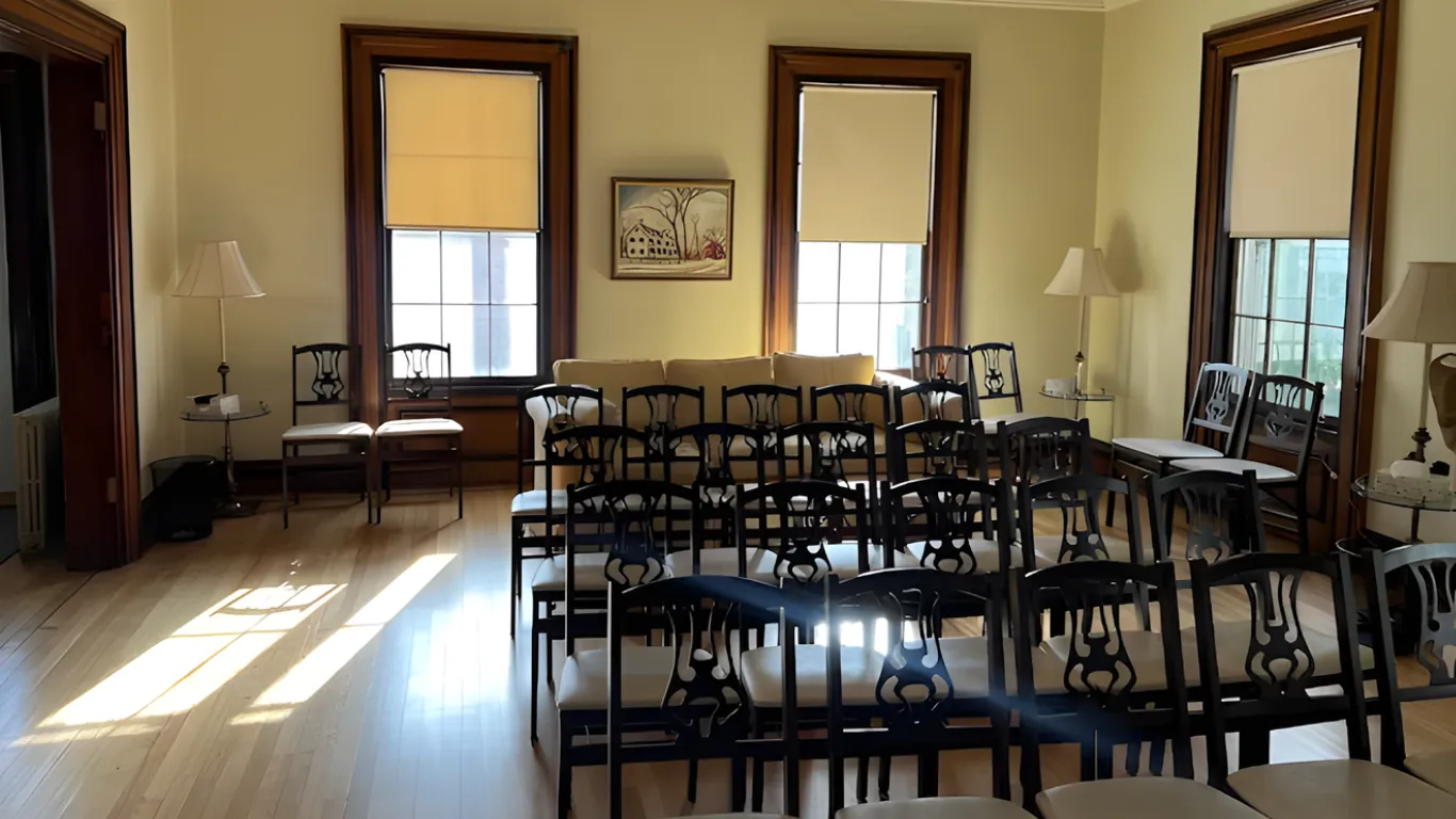 Empty meeting room with rows of black chairs, tall windows, and sunlight streaming across wooden floors