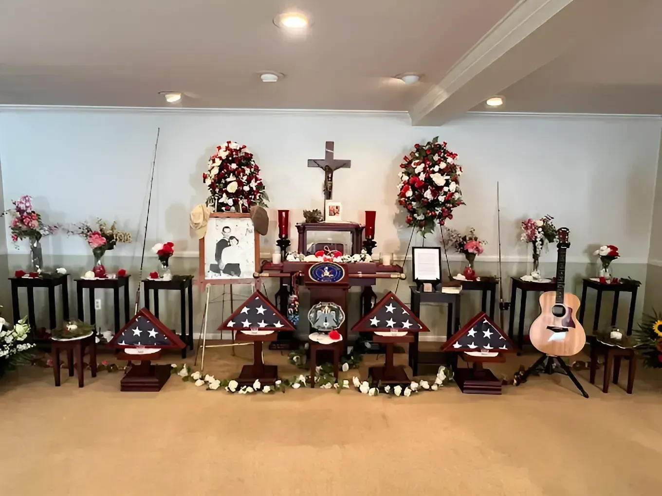 Memorial altar with flowers, candles, crosses, framed photos, and guitars in a decorated room.