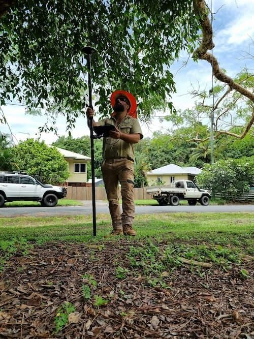 A Man Is Standing Under a Tree Holding a Pole and A Tablet — Advanced Vegetation Management in Kewarra Beach, QLD