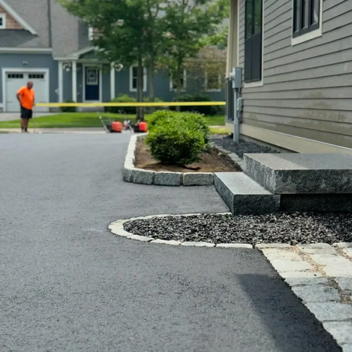 Asphalt driveway with stone steps and edging, bushes, and a person in orange shirt standing in a residential area.