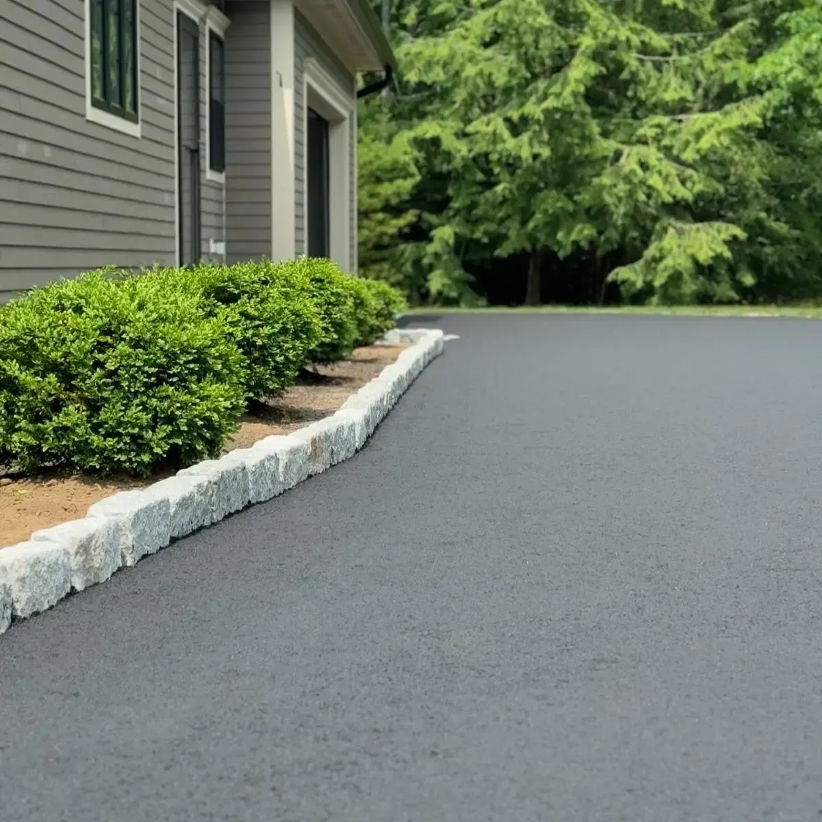 Asphalt driveway with stone edging and green bushes, next to a gray house with a garage.