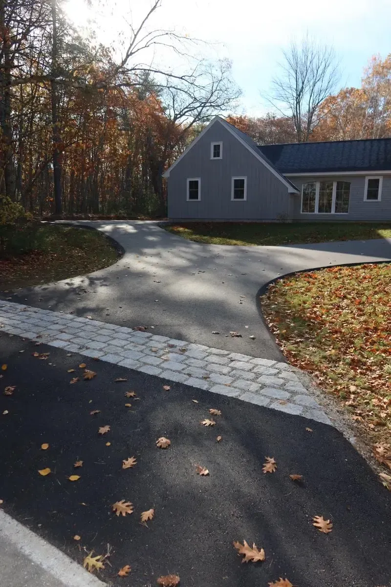 Driveway curves towards a gray house with a black roof, surrounded by fall foliage.