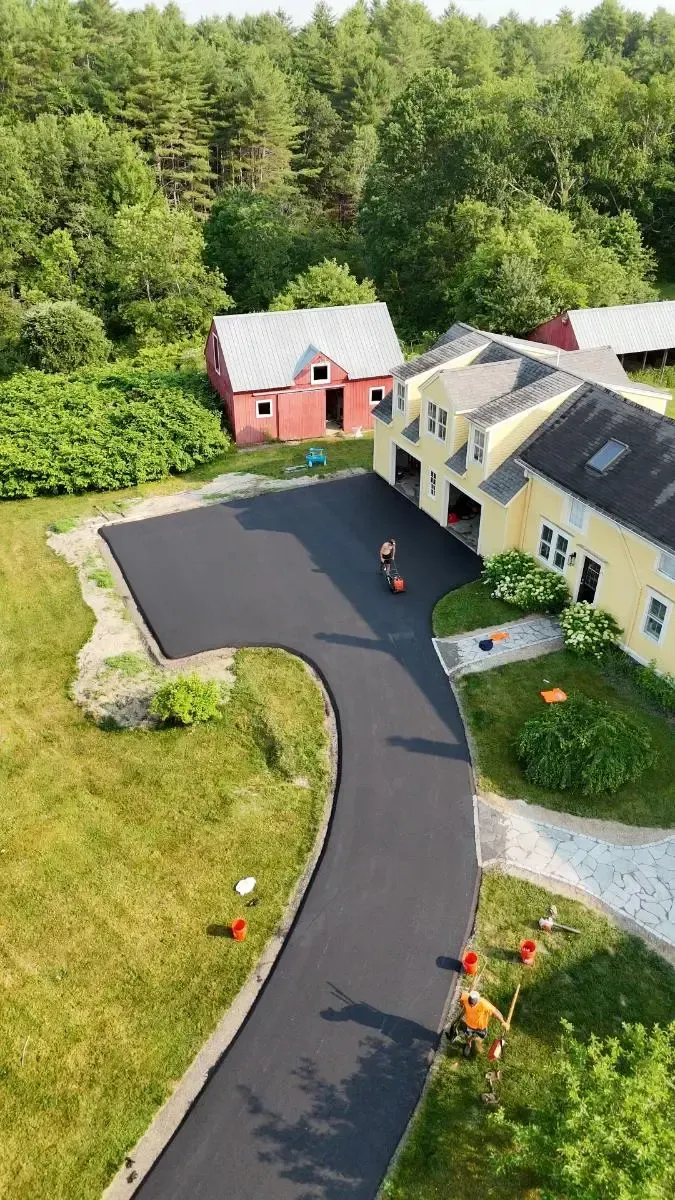 Newly paved driveway curves towards a yellow house, red barn in the background. Green trees and grass surround.