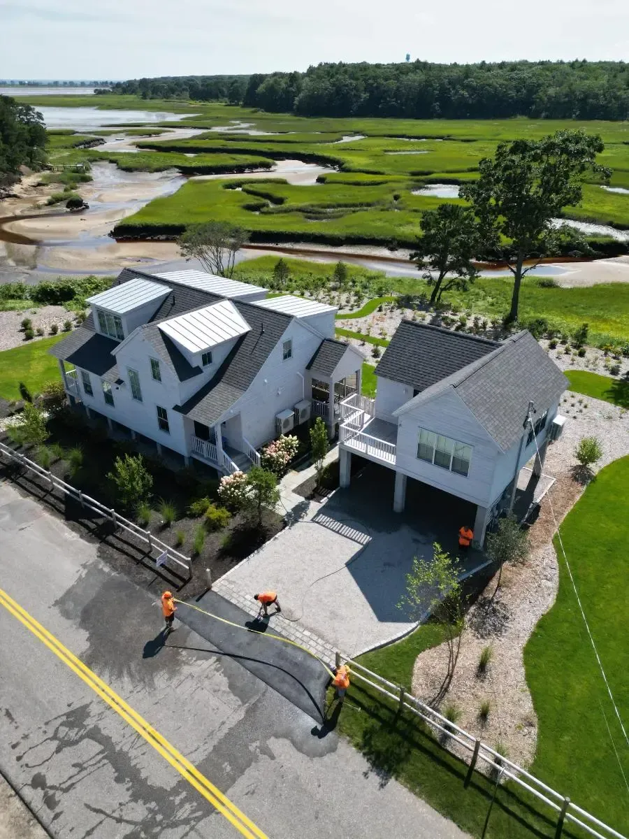 Two white houses with metal roofs, next to a marsh, workers paving a driveway.
