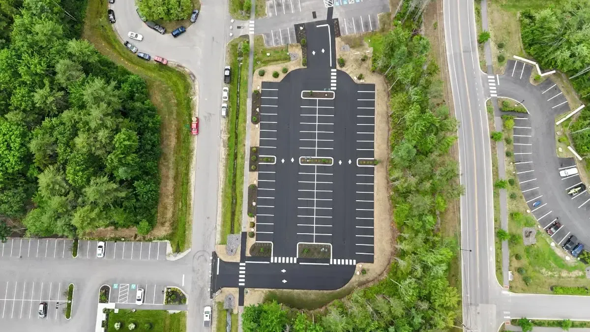 Overhead view of a new asphalt parking lot surrounded by trees, with a road to the right and other parking areas nearby.