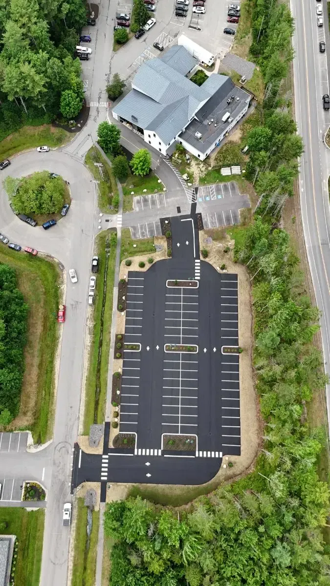 Aerial view of a parking lot and buildings; black asphalt, white parking lines, green trees, gray roofs, and a roundabout.
